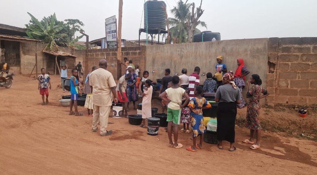 Local partner kneeling at a newly installed water well pump surrounded by community members waiting to draw water in a rural village.